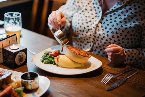 A woman pouring gravy on a plate containing The Best Ever Pub Pie, potato and vegetables