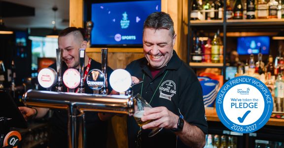 Two team members behind a bar with the Dyslexia Friendly Workplace pledge badge in corner