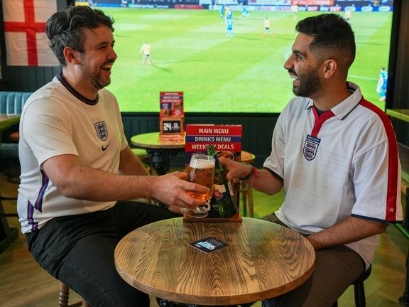 Two people sitting at a pub table with football being shown on a big screen in the background